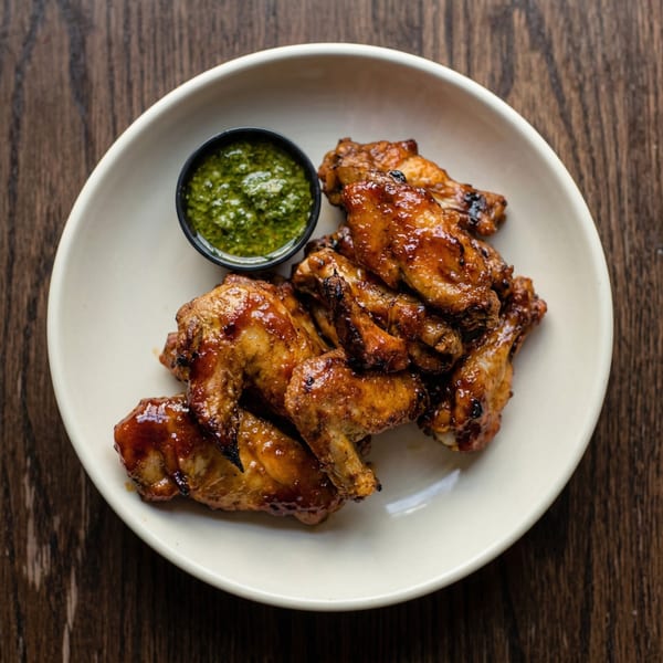 A white bowl filled with glazed chicken wings and a small cup of green dipping sauce.