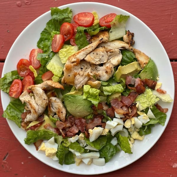 A vibrant Cobb salad with grilled chicken, bacon, avocado, tomatoes, and hard-boiled eggs on a white plate.