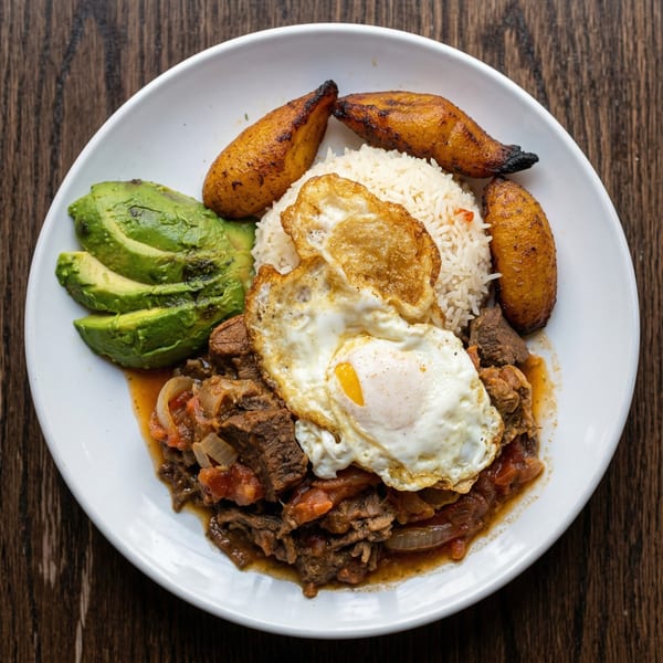A white plate with a meal of stewed beef, rice, fried eggs, avocado slices, and plantains.