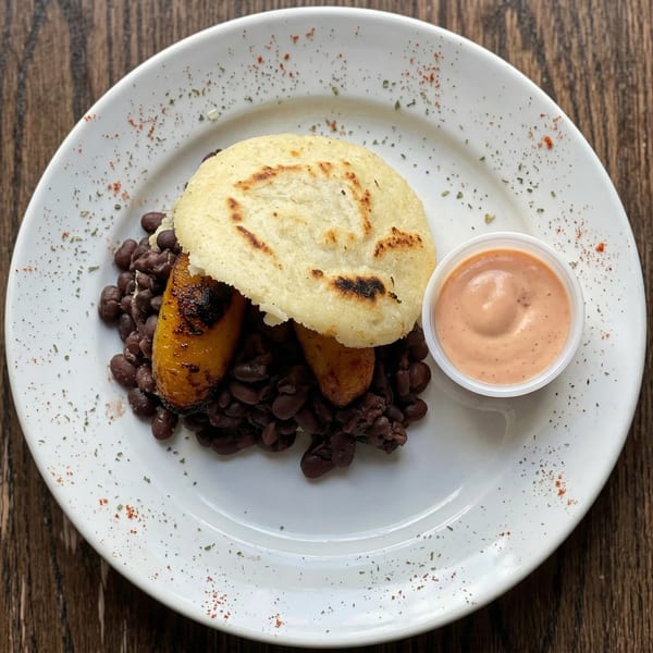 Arepa, black beans, and plantains with a side of pink sauce on a white plate.